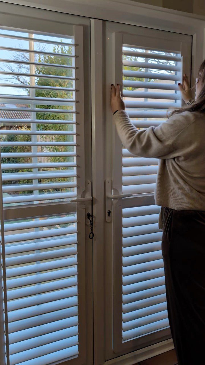 A person adjusting white plantation shutters on a set of glass patio doors.