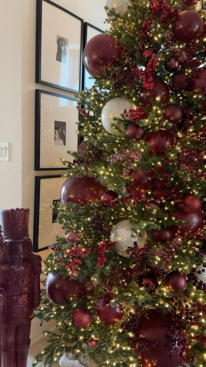 Decorated Christmas tree with red and burgundy ornaments, berries, and warm glowing lights against a wall.