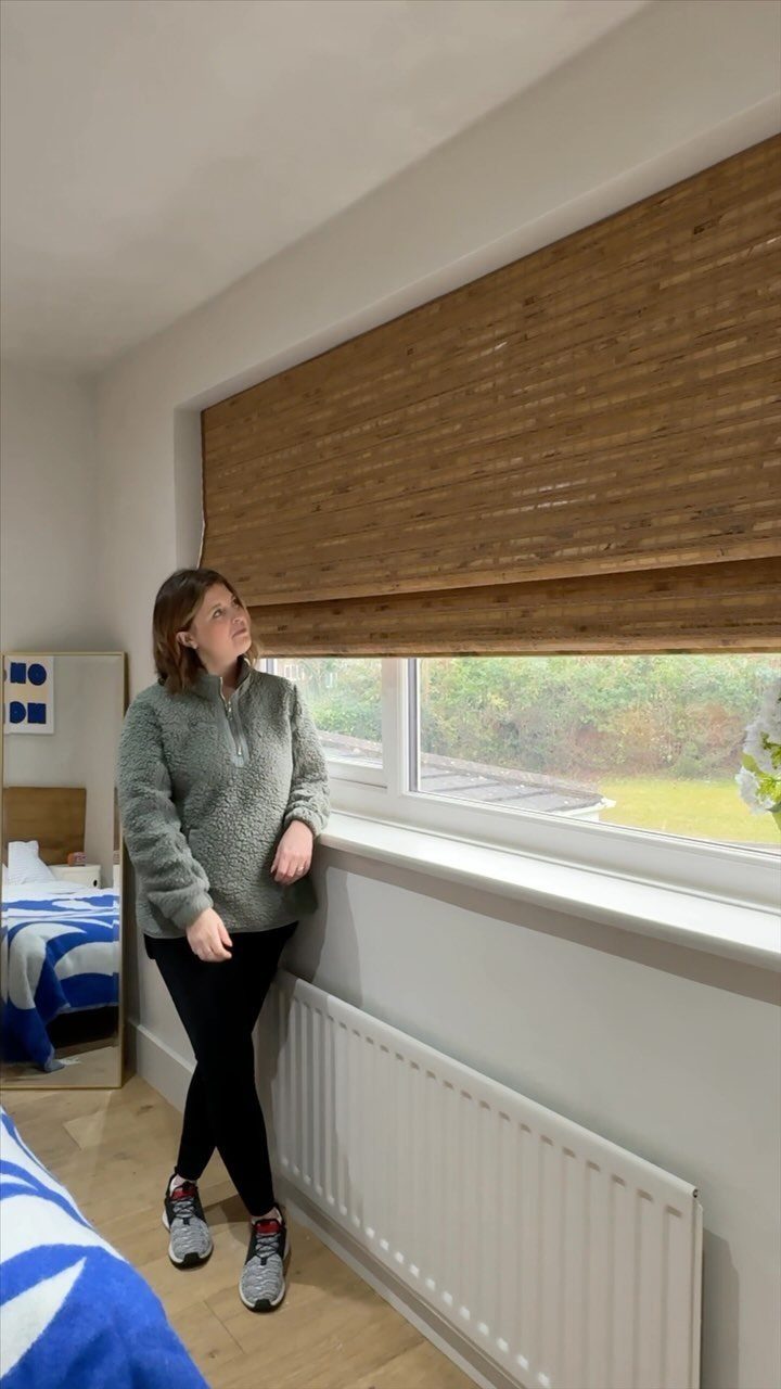 A woman standing by a window with a textured bamboo roman blind installed.