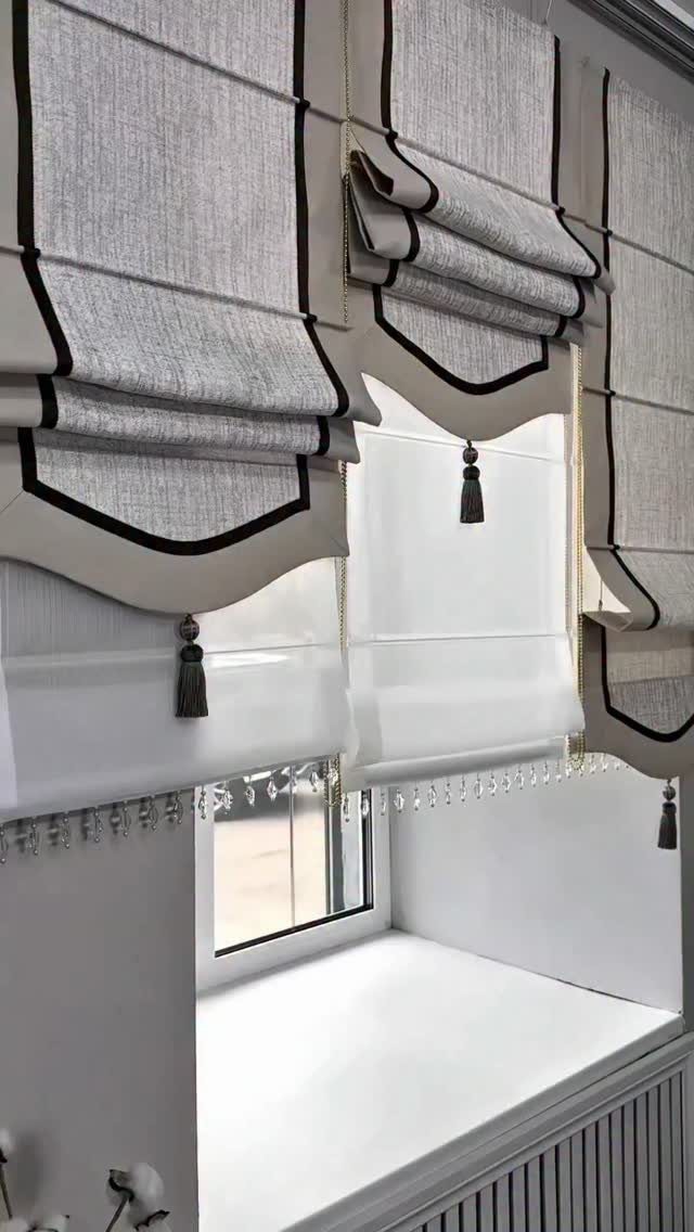 Light grey roman blinds with black piping and tassels hanging over a white window.