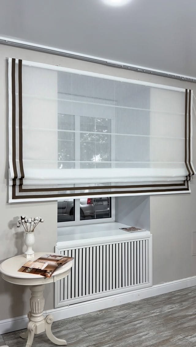 A white roman blind with dark brown side stripes hanging over a window above a radiator.