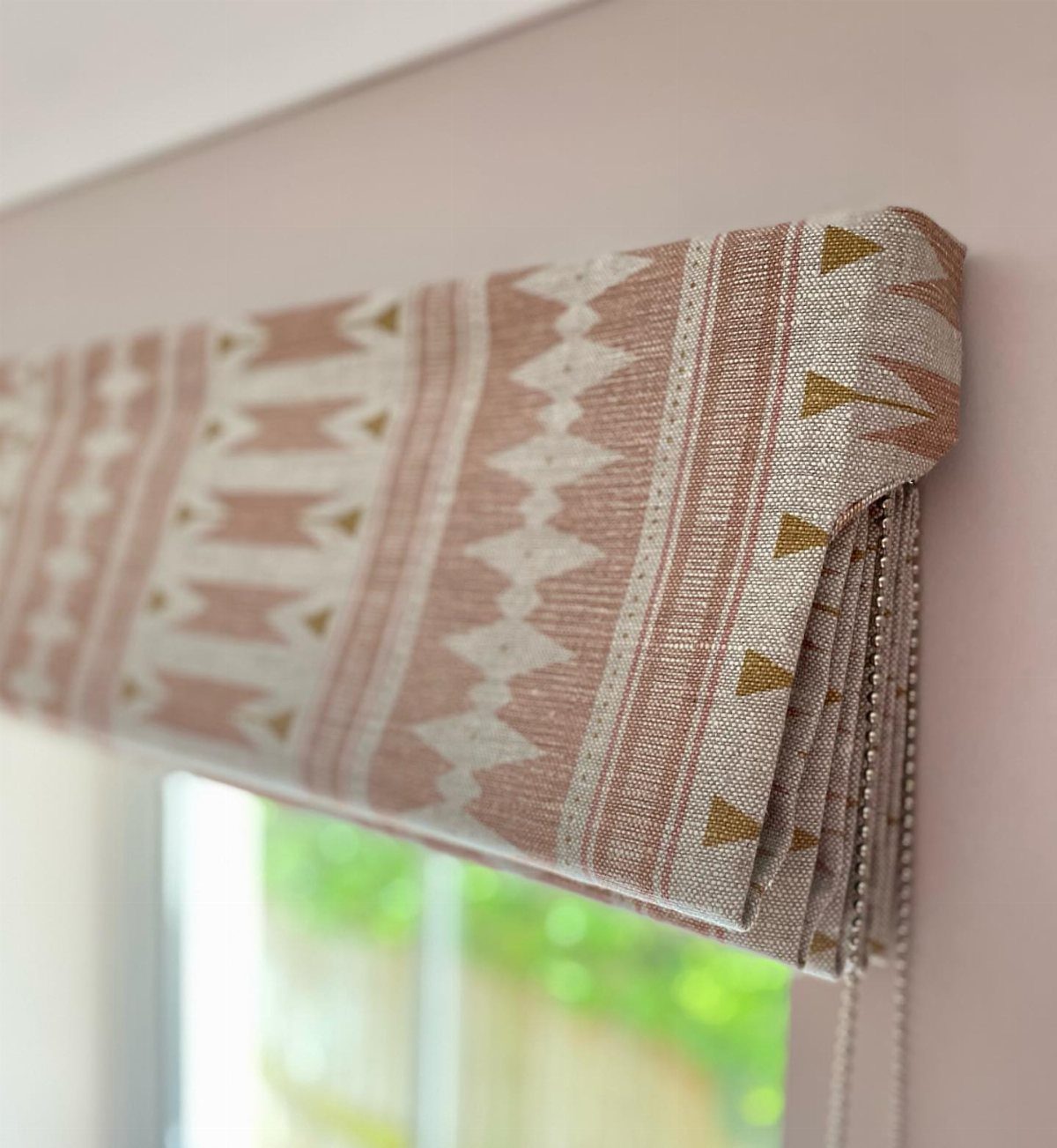 A patterned roman blind with geometric shapes in rose and cream tones hanging over a window.