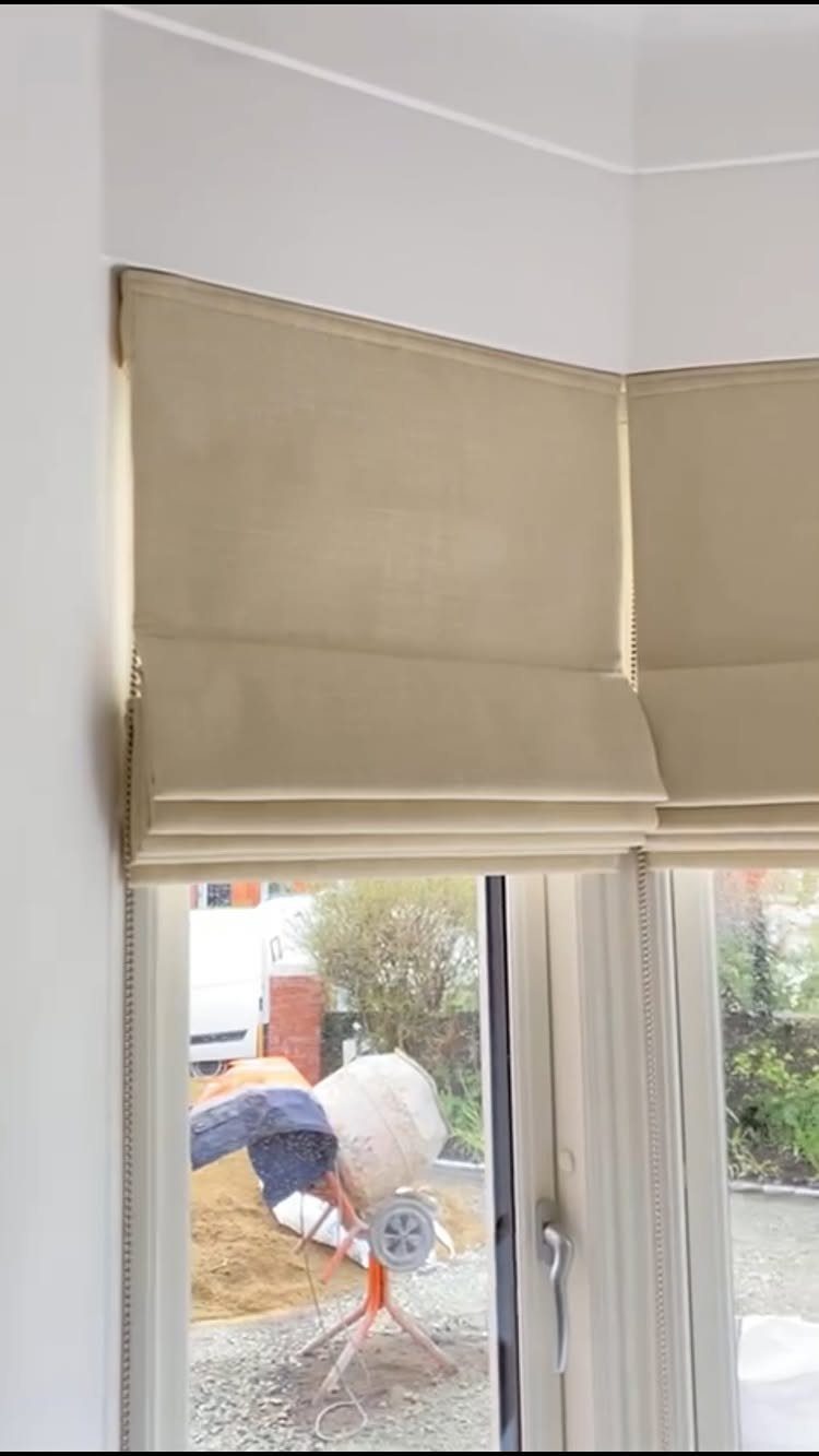 Beige fabric roman blinds installed on a white bay window overlooking an outdoor garden area.