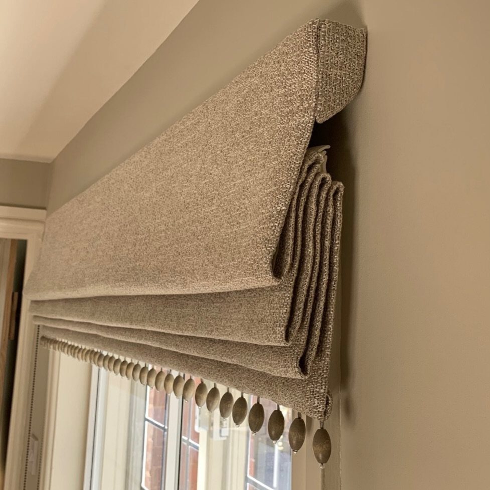 A textured grey fabric roman blind with decorative bead trimmings hanging over a window.