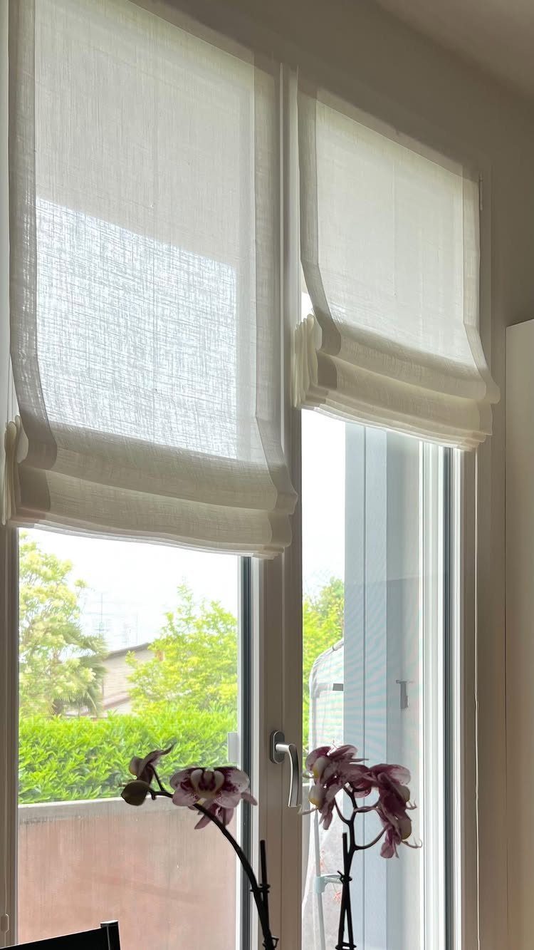 Two white textured roman blinds partially raised on a tall window overlooking green trees.