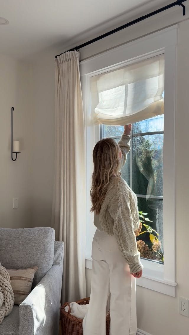 Roman Shades with Curtains: 8 Looks That Work 1 Woman adjusting a cream roman shade on a window framed by long curtains.