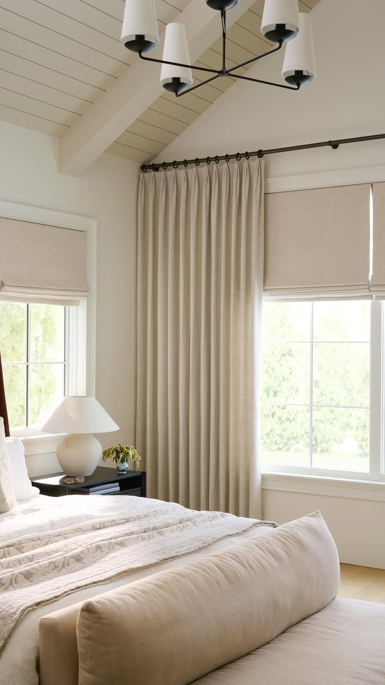 Roman Shades with Curtains: 8 Looks That Work 8 Beige roman shades paired with long floor-length curtains on windows in a bright bedroom.