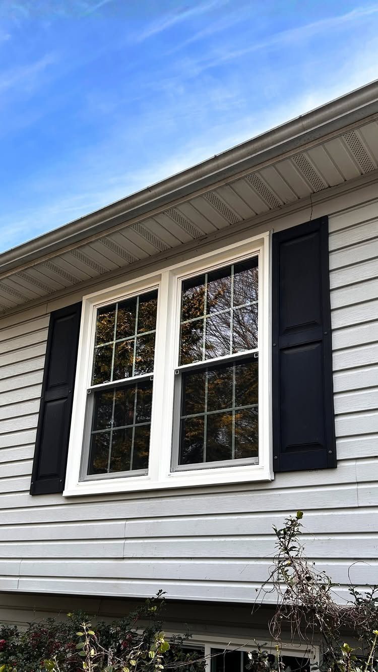 Two white double-hung windows with black shutters on grey siding under a blue sky.