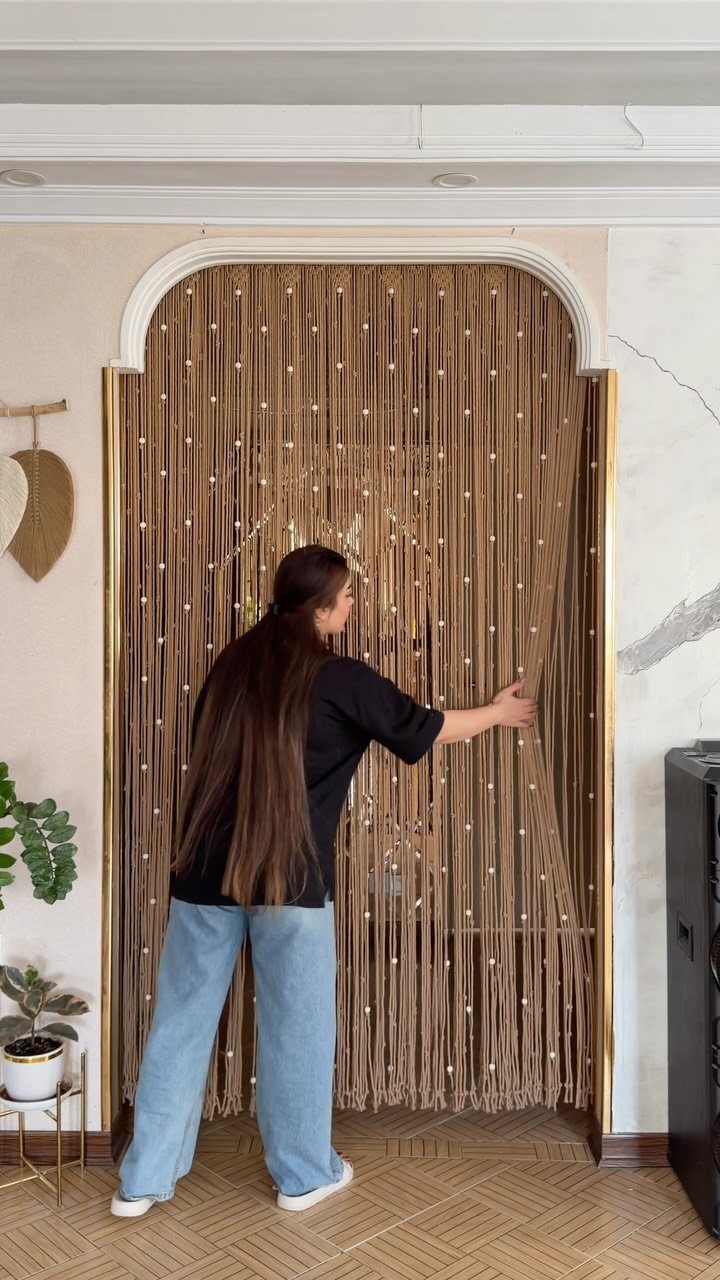 11 Ways to Style Curtains for Every Room in 2026 8 A woman touches a macrame string curtain with white beads hanging in an arched doorway.