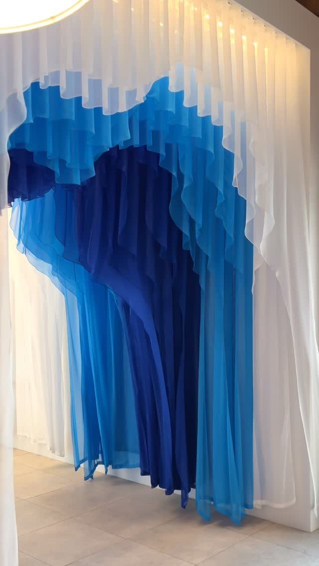 11 Ways to Style Curtains for Every Room in 2026 10 Layered blue and white sheer curtains hanging in a wavy, sculptural pattern against a white wall.