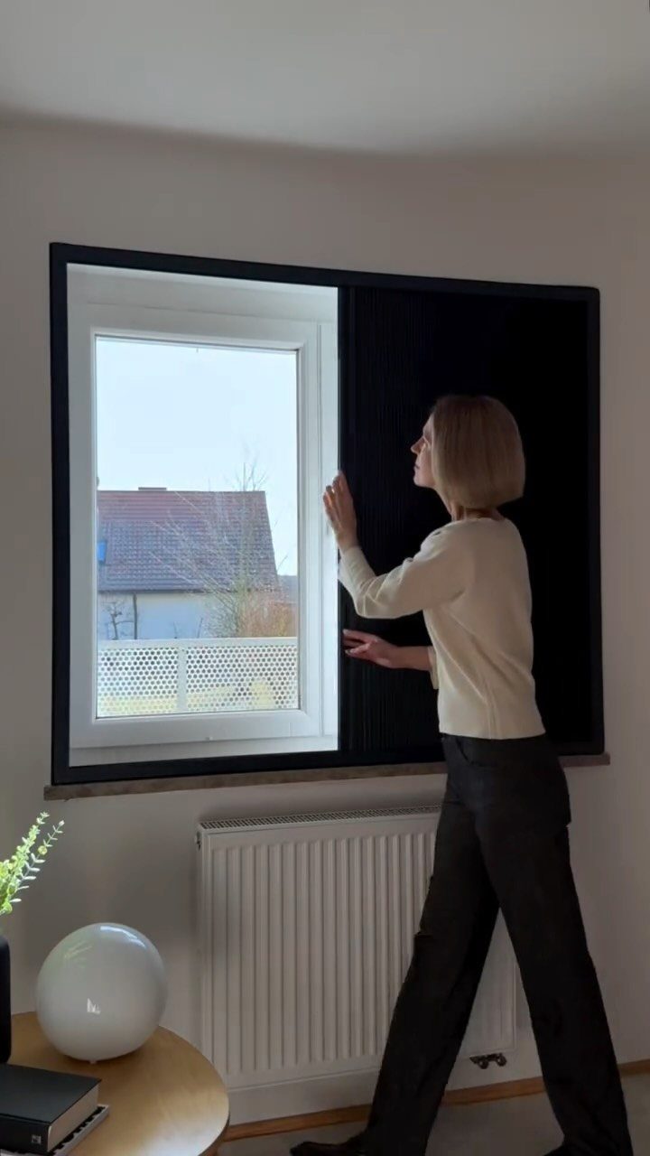 Window Blinds for Living Room: 11 Photo Ideas 7 A woman opening a large black blackout blind to reveal a view of a house outside.