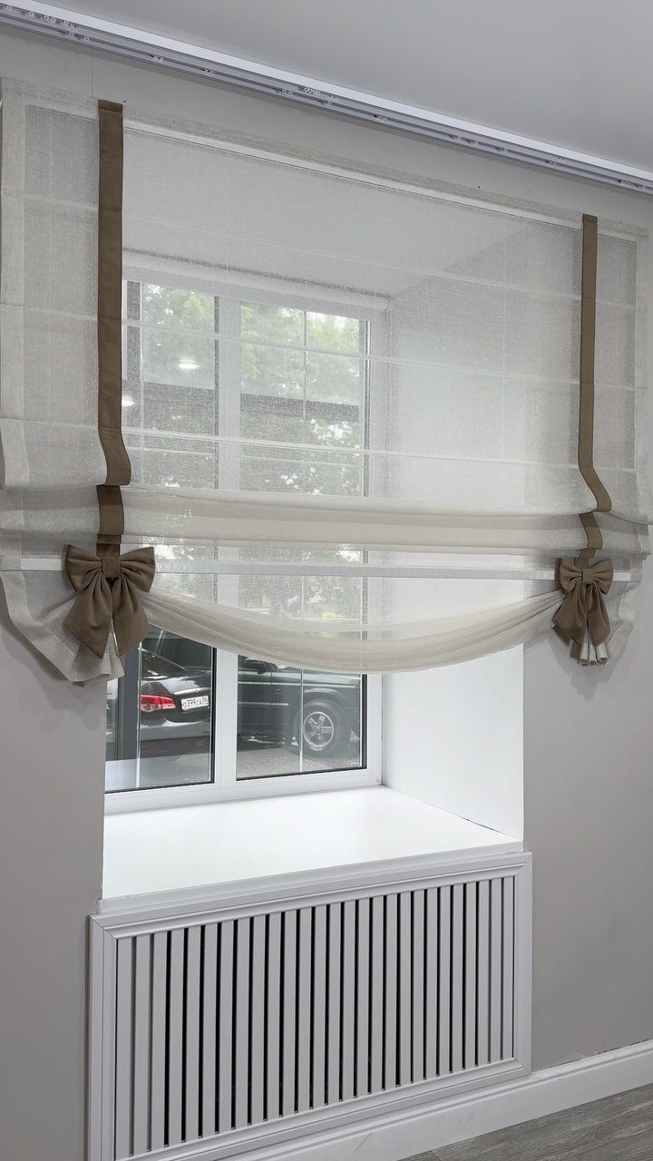 Window Blinds for Living Room: 11 Photo Ideas 1 White fabric Roman shades with tan ribbon bows hanging over a large window.