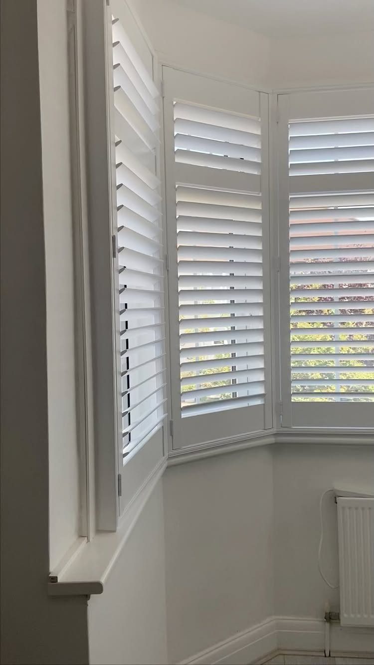 Window Blinds for Living Room: 11 Photo Ideas 2 White plantation shutters installed on a large bay window in a bright room.
