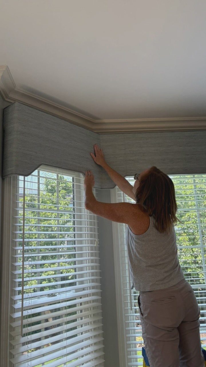 Window Cornice DIY: 5 Ways With Fabric, Wood, and Molding 5 A woman adjusting a grey fabric window cornice installed above white blinds.