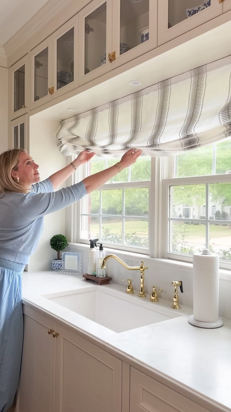 10 Window Dressing Ideas for Shutters, Shades and Blinds 4 A woman adjusting a patterned fabric Roman shade over a white kitchen window.