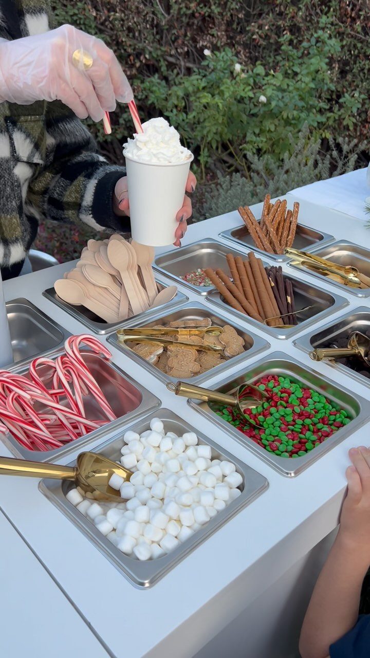 A person holds a cup of hot cocoa with whipped cream near a hot chocolate bar buffet.