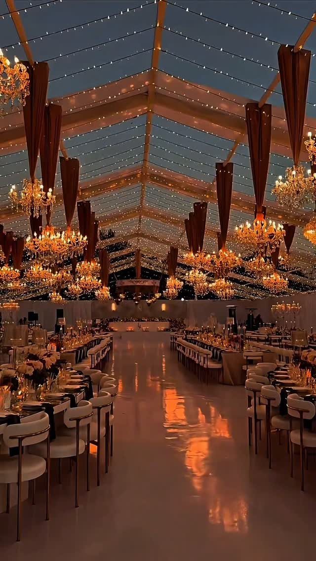 Warm chandeliers and twinkling lights hanging from a transparent marquee tent over wedding reception tables.